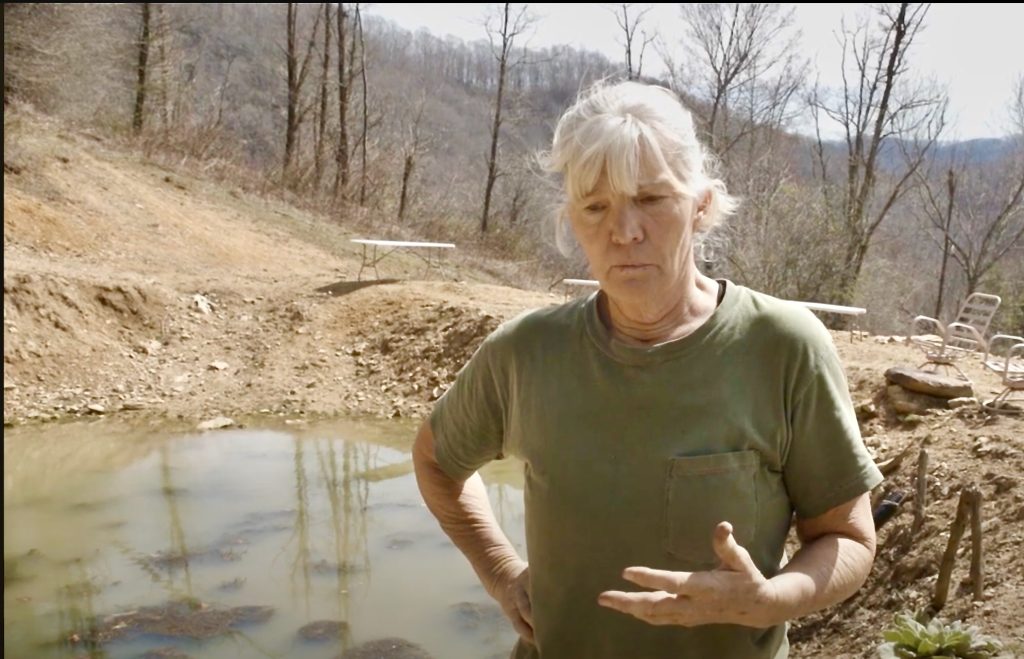 A sad looking woman with downcast eyes stands beside a pond that is nearly dried up.